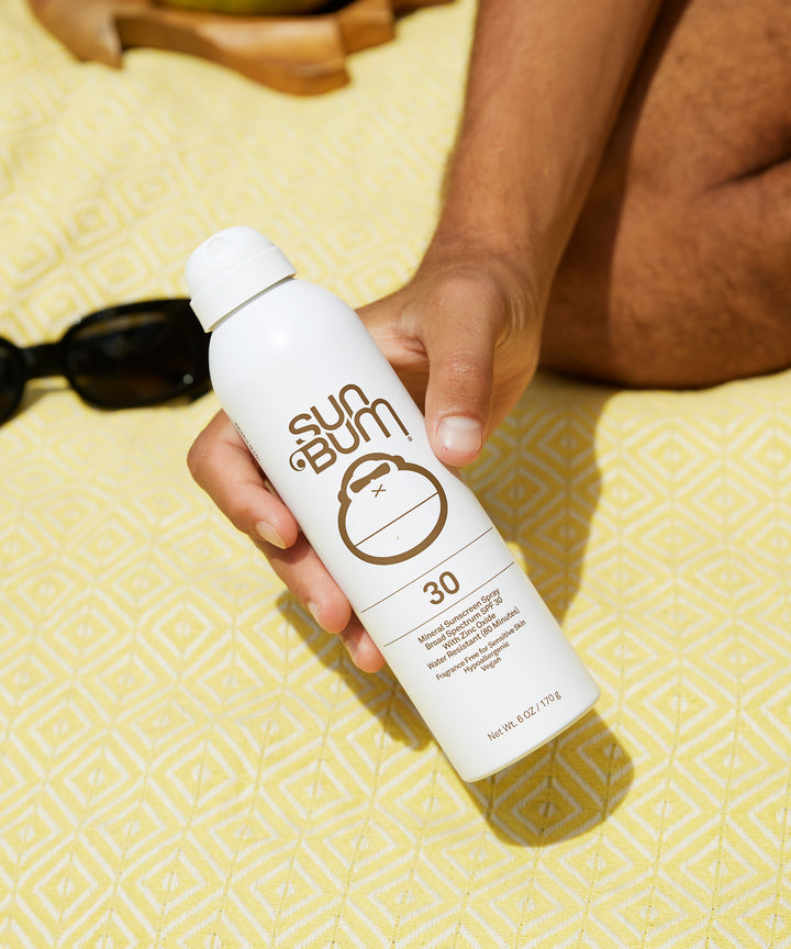 Hand holding a bottle of Mineral SPF 30 Sunscreen Spray on a yellow patterned blanket with sunglasses in the background, Sun Bum