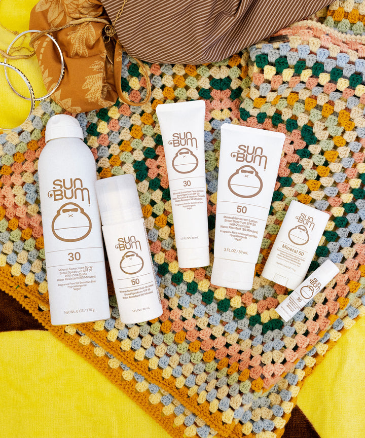 A collection of Sun Bum Mineral SPF 30 and SPF 50 sunscreens arranged on a colorful crochet blanket, showcasing various product sizes and packaging options. Sun Bum