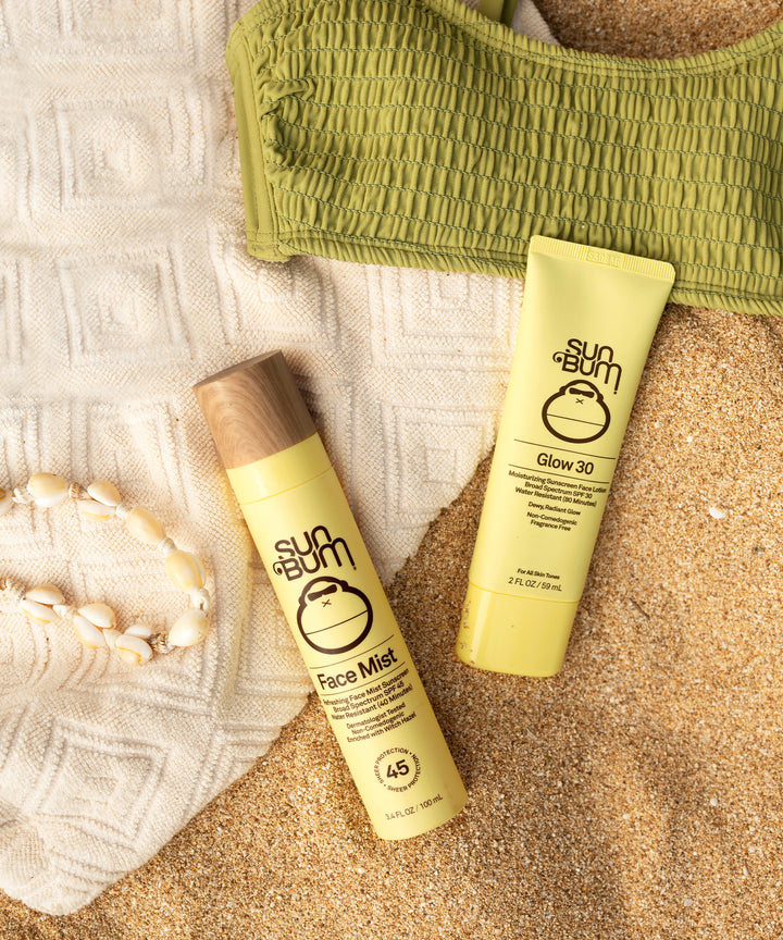 SPF 45 Sunscreen Face Mist by Sun Bum on sandy beach with SPF 30 sunscreen and green swimsuit top nearby, lightweight protection for sunny days.