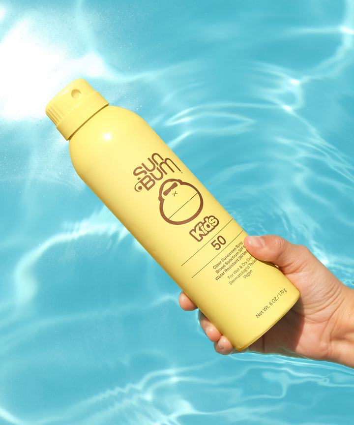 Hand holding a yellow bottle of Kids SPF 50 Clear Sunscreen Spray against a blue water background. Sun Bum.