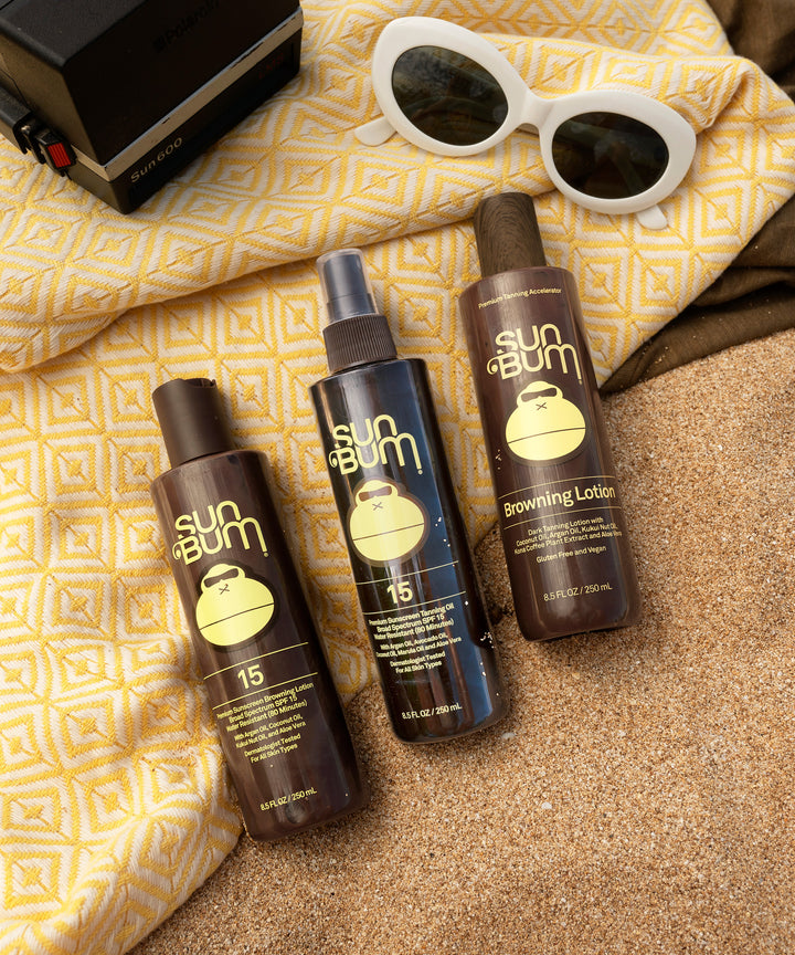 Three bottles of SPF 15 Sunscreen Tanning Oil and Browning Lotion on a sandy beach with a yellow patterned towel, white sunglasses, and a vintage camera, ideal for achieving a golden tan while keeping skin smooth and protected, Sun Bum.