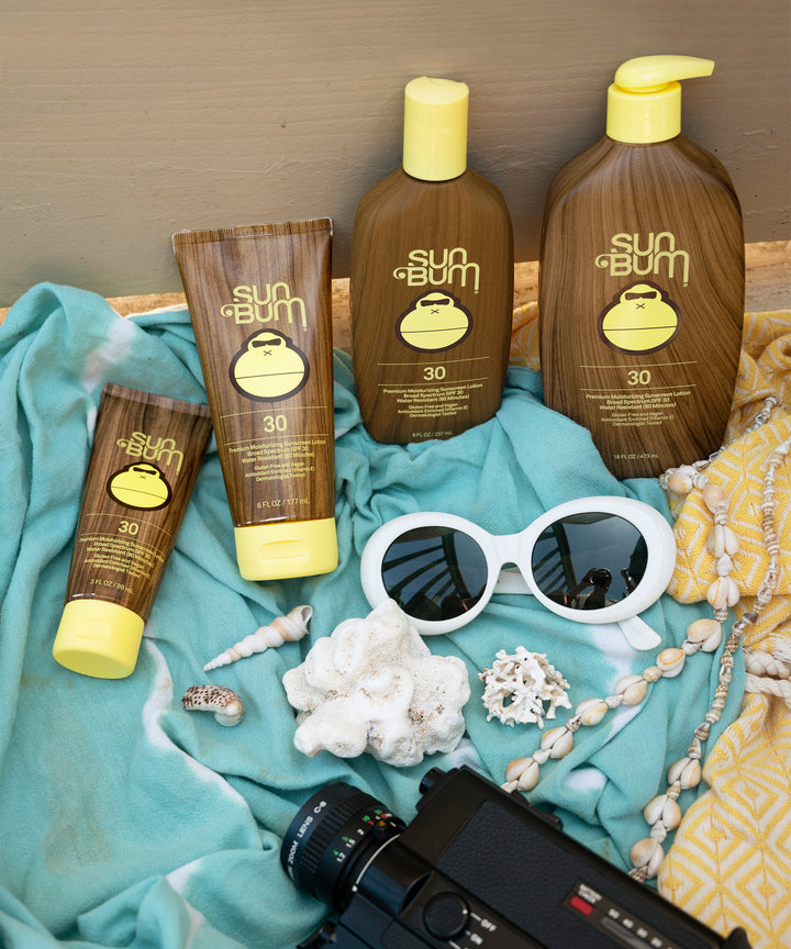 Bottles of Original SPF 30 Sunscreen Lotion by Sun Bum arranged on a beach towel with sunglasses, seashells, and a camera.