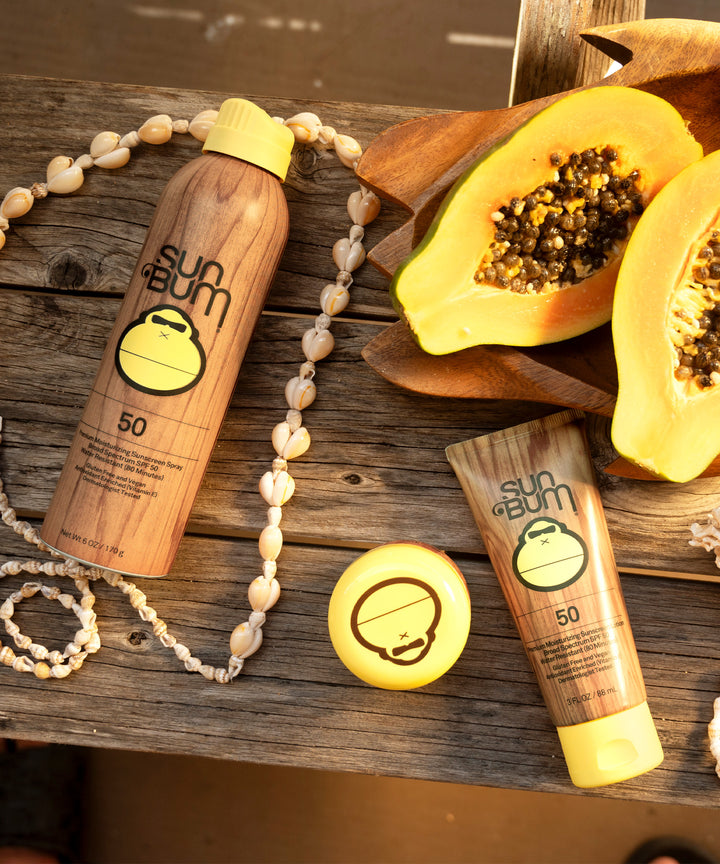 Sun Bum Original SPF 50 Sunscreen Spray placed on a wooden surface with sliced papaya and shell necklace.