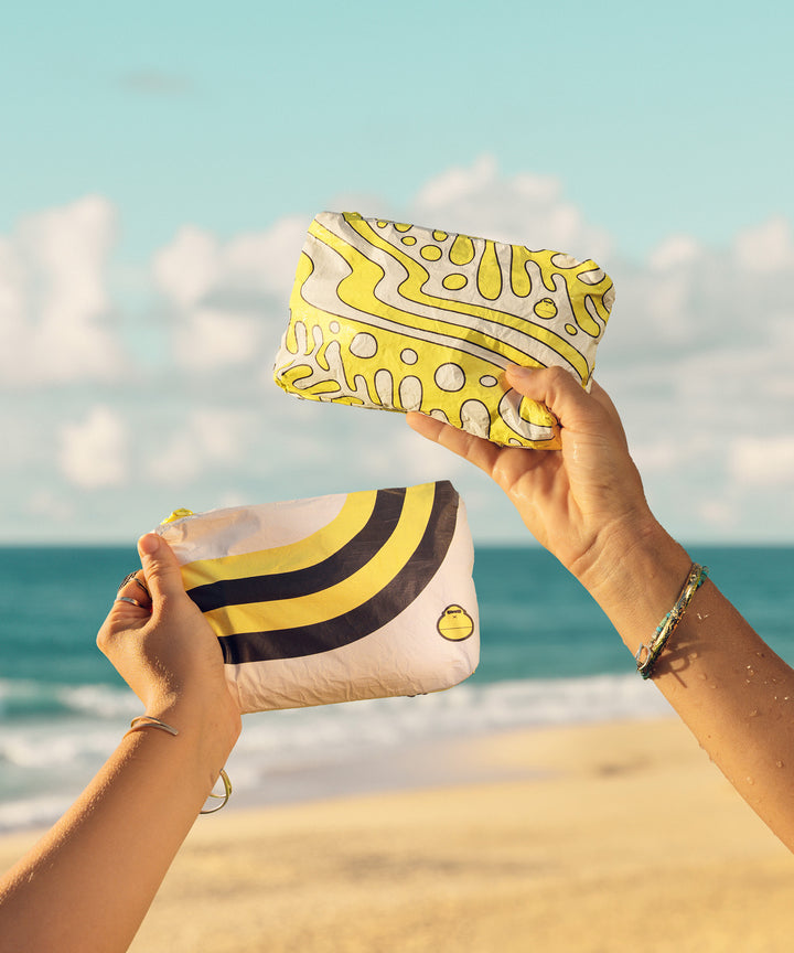 Two hands holding splash-proof pouches for the Hair Care Travel Essentials Kit against a beach backdrop; designed in collaboration with ALOHA Collection. Sun Bum