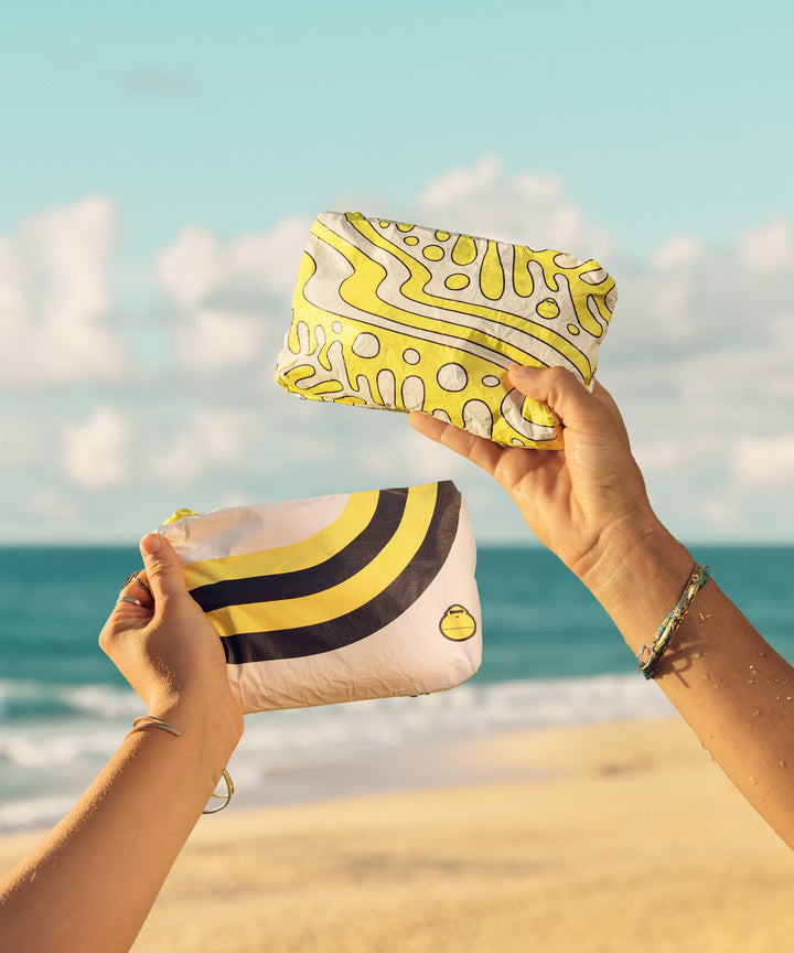 Two hands holding up a yellow and white splash-proof pouch with wavy designs at a beach, perfect for storing your skin care travel essentials from Sun Bum.