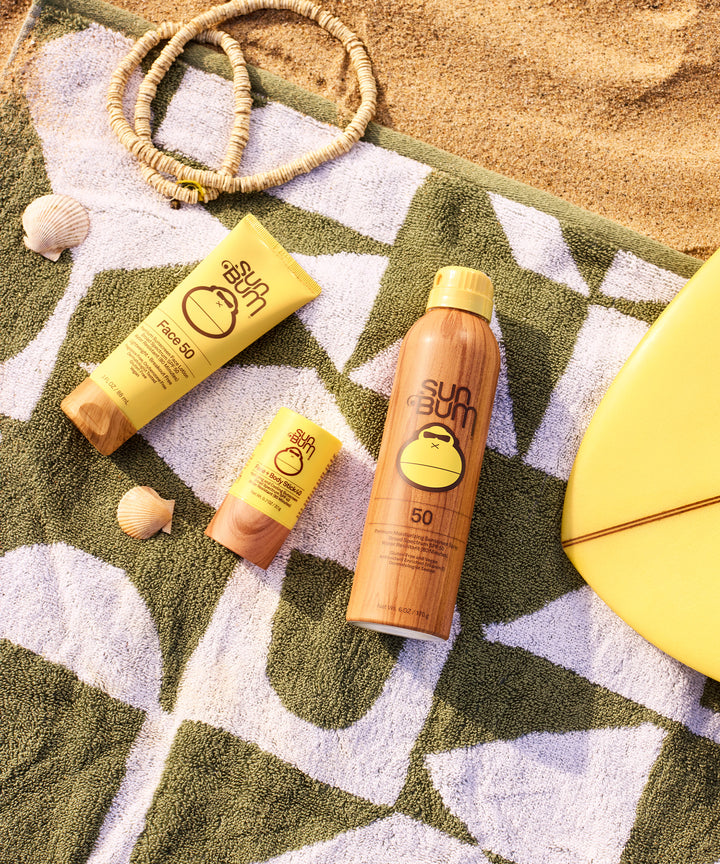 Sun protection essentials on beach towel: SPF products, including Original Face and Body Stick, arranged with seashells and beaded necklace. Perfect for effortless, on-the-go sun care. Sun Bum