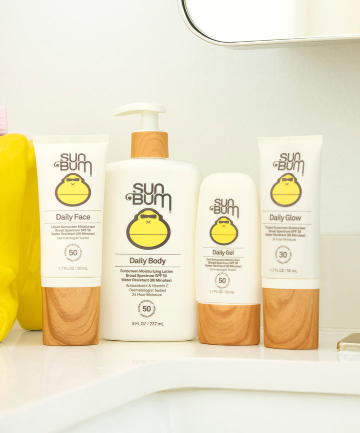 Sun Bum skincare products displayed on bathroom counter, including Daily Face, Daily Body, Daily Set, and Daily Glow moisturizers with SPF protection. Sleek packaging with iconic Sun Bum logo. Sun Bum