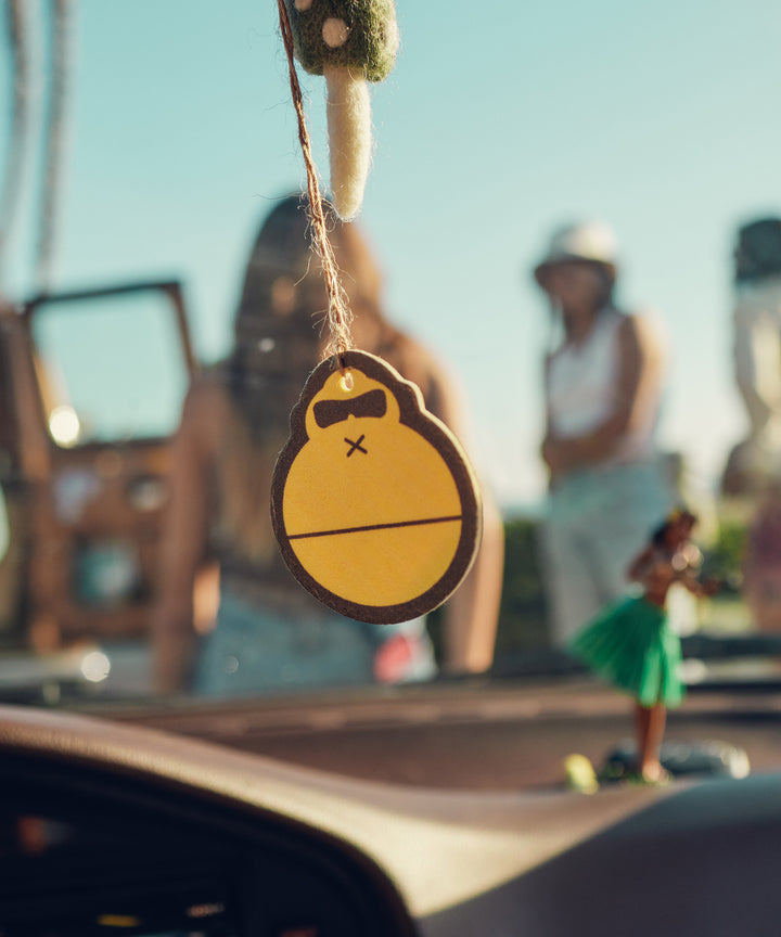 A cheerful cartoon air freshener hanging from a car's rear-view mirror, designed to combat car odors and bring a fresh, summery vibe. Sun Bum