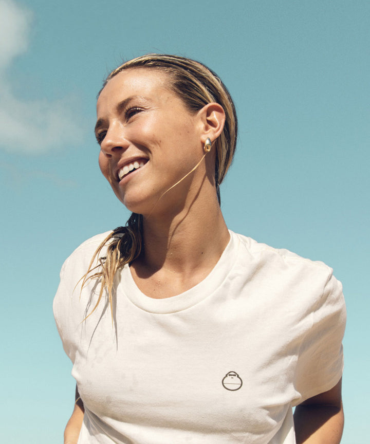 Woman smiling, wearing a white plain tee with a small mascot logo under sunny blue sky, Sun Bum.