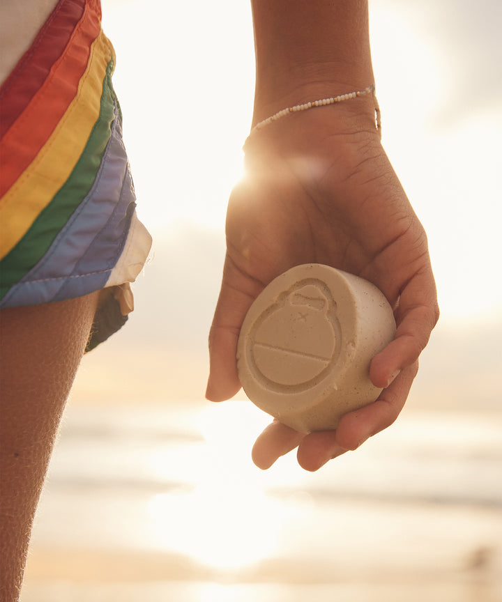 Person holding surf wax near ocean, ready for use, Sun Bum.