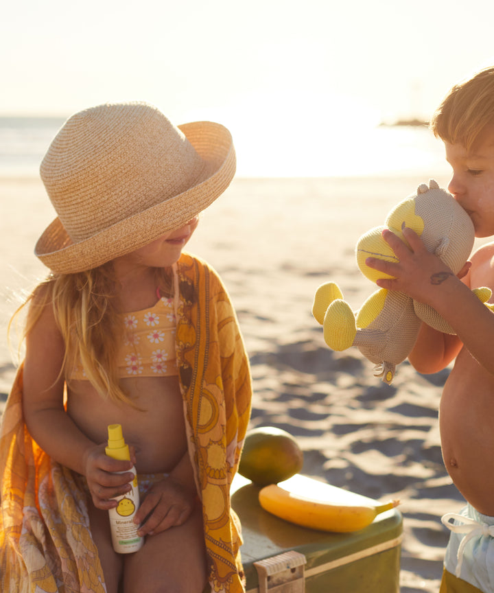 Two kids playing on the beach, one holding a bottle of Baby Bum Mineral SPF 50 Sunscreen Spray, Baby Bum.